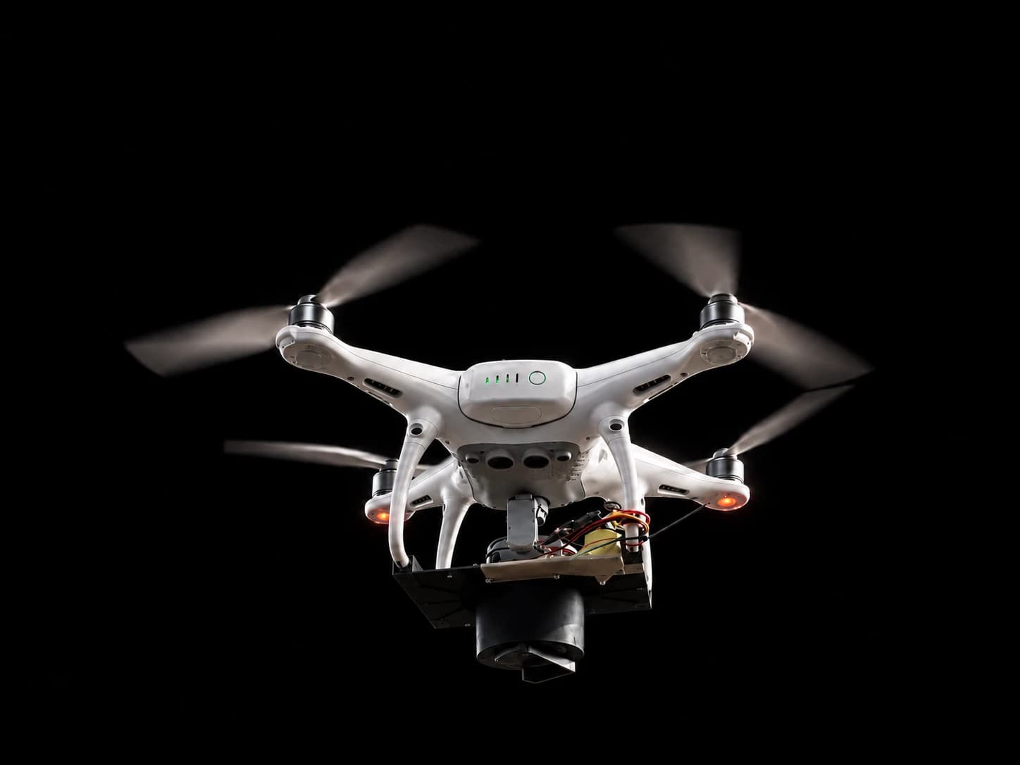 A white quadcopter in flight against black, carrying a custom seed-dispersal payload: wired electronics on a black mounting plate above a cylindrical drop tube.